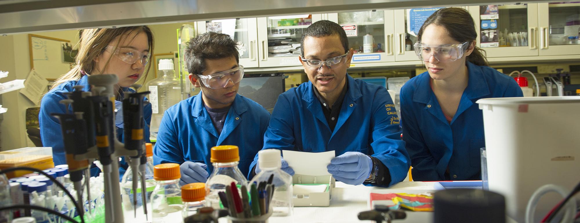 A group of four people in blue lab coats and protective eyewear in a laboratory setting examining a small white sheet.
