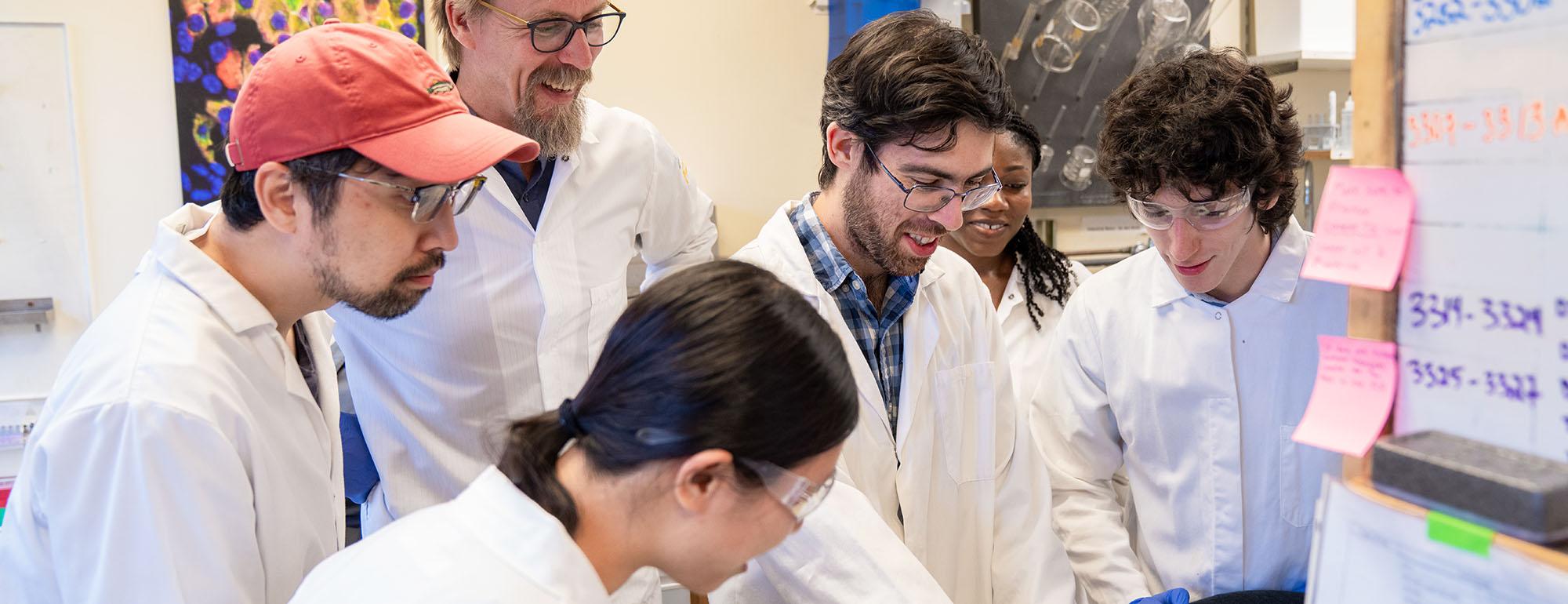 Researchers in lab coats gather around a bench, smiling and reviewing notes together in a brightly lit laboratory