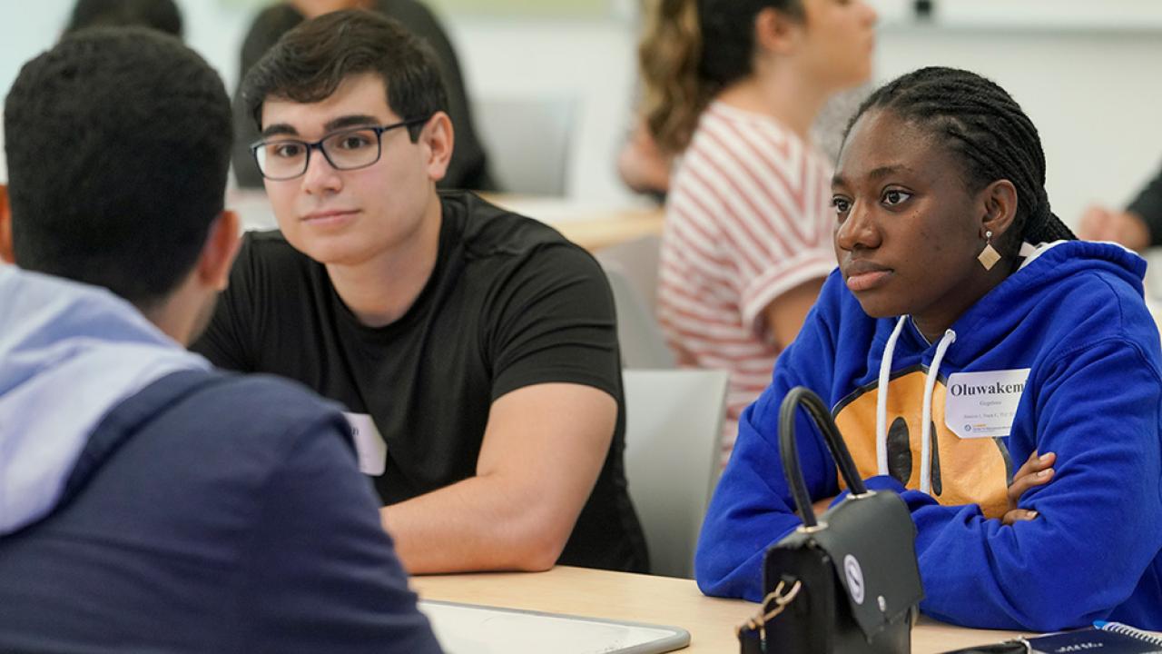 A group of male and female students sitting at a table listening to a speaker