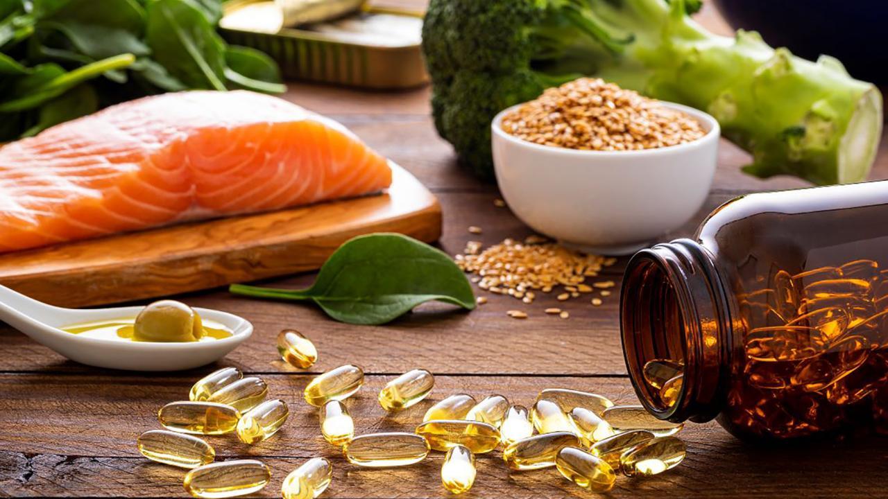alt="A close-up of nutrient-rich foods and supplements on a wooden surface, featuring a raw salmon fillet on a cutting board, a bowl of sesame seeds, fresh spinach leaves, broccoli, and a white spoon containing olive oil and an olive. Nearby, an amber bottle is tipped over, spilling golden omega-3 capsules."