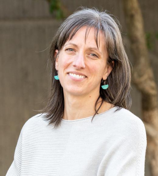 A woman with shoulder-length gray hair, wearing turquoise earrings and a light-colored sweater, smiles at the camera with a blurred outdoor background.