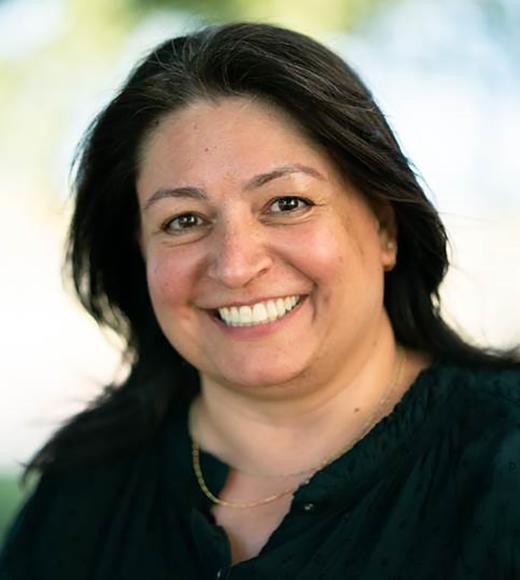 The image features an individual with long dark hair wearing a black top and a gold necklace. The background appears to be outdoors with blurred greenery, suggesting a shallow depth of field typically used in portrait photography.