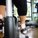 Close-up of a person’s legs pedaling on a stationary exercise bike in a gym, with another individual running on a treadmill in the background.