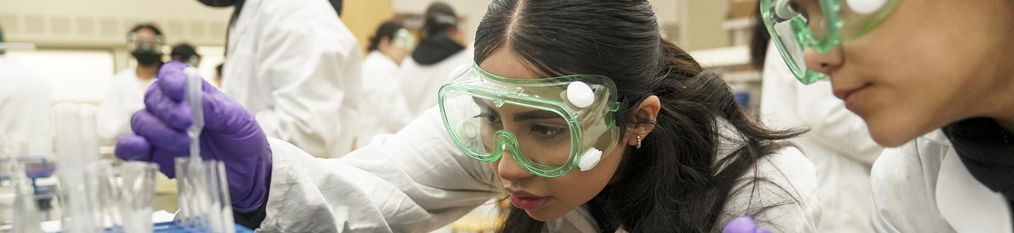Two female students in PPE with a test beaker and dropper in a lab class